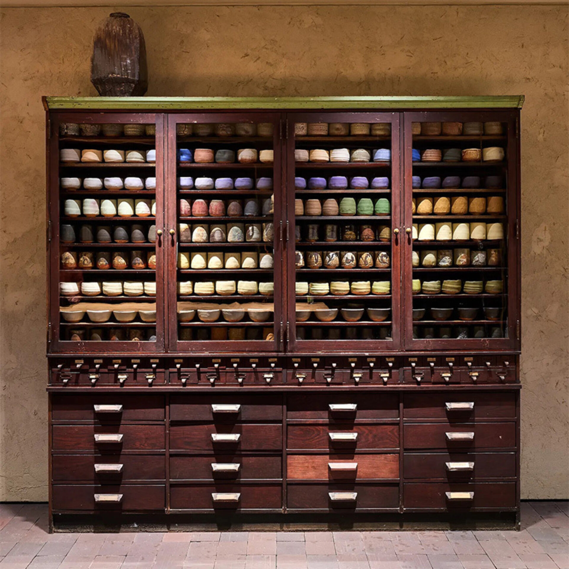 Wooden apothecary cabinet with glass doors showing rows of stacked colorful ceramic bowls on shelves above labeled drawers and jar on top.