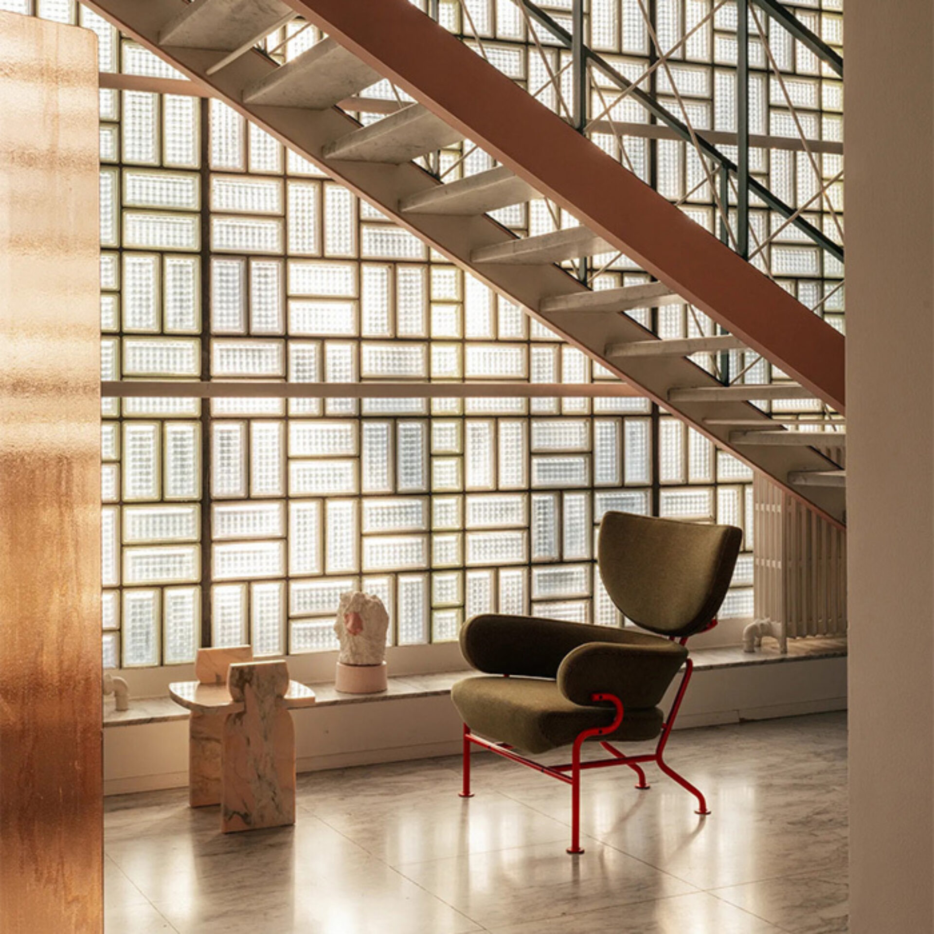 Sunlit interior with a diagonal metal staircase over a glass-block wall, featuring an olive-green lounge chair with a red frame, marble side table and small sculptures on the ledge.