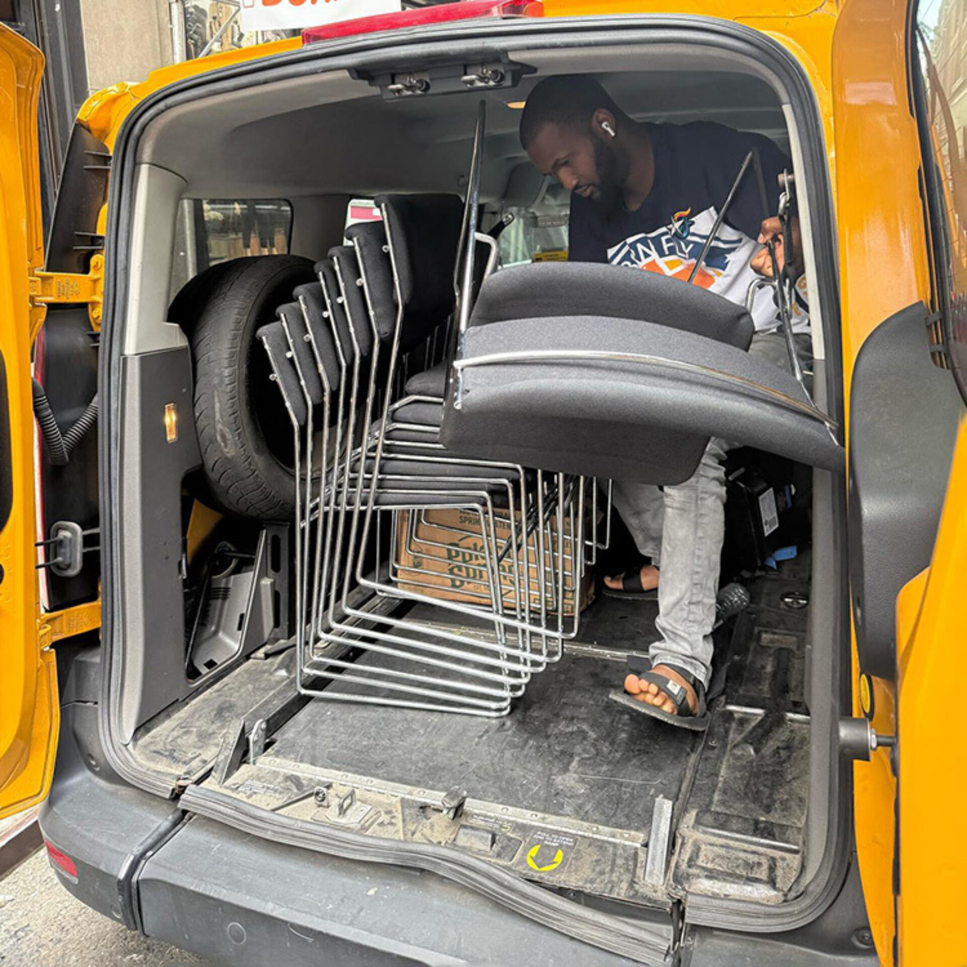 A man loads multiple stacked chairs and a spare tire into the back of a yellow taxi, surrounded by packing boxes and bags.