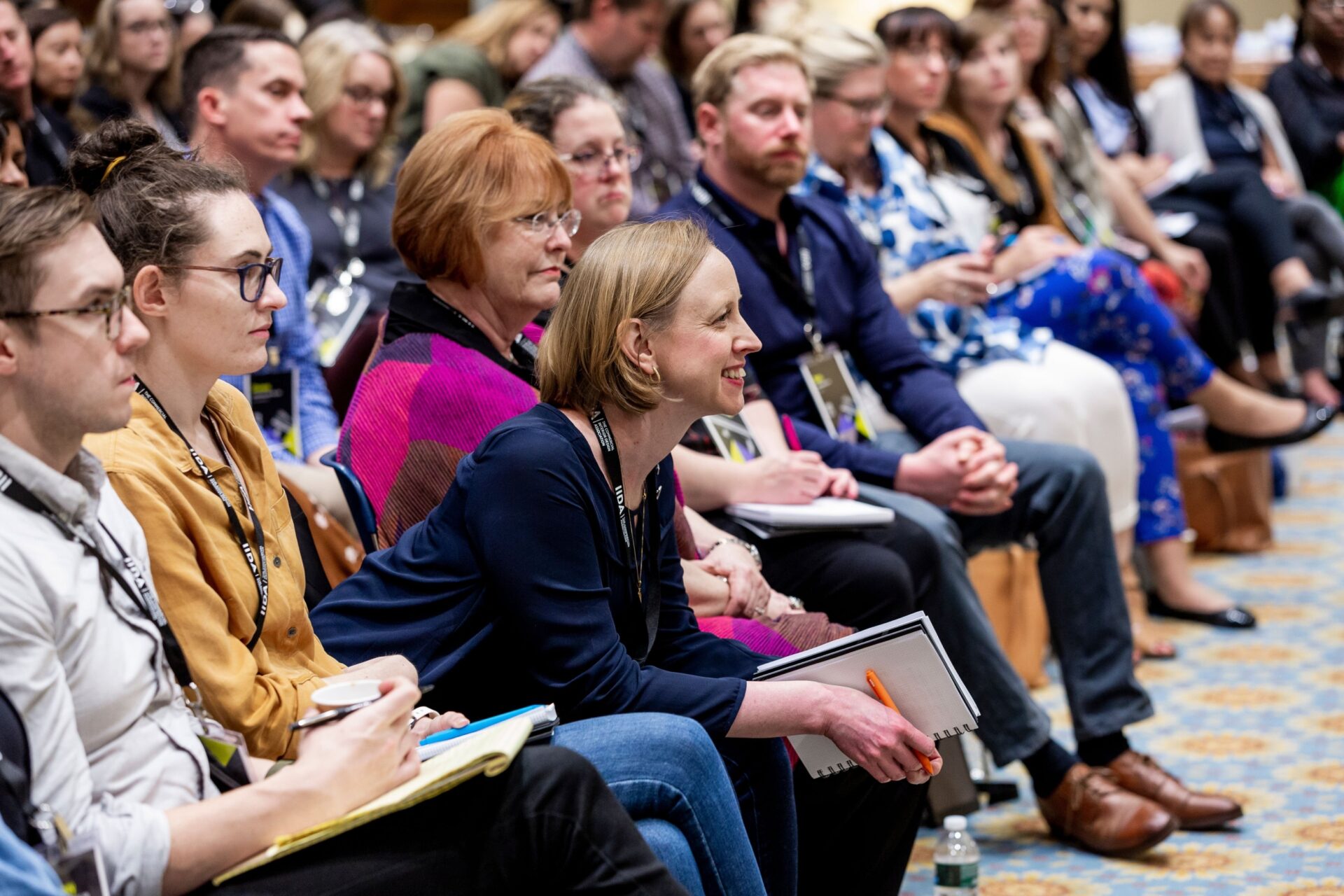 A diverse group of attendees focuses intently on a speaker during a conference, with many taking notes and participating actively.
