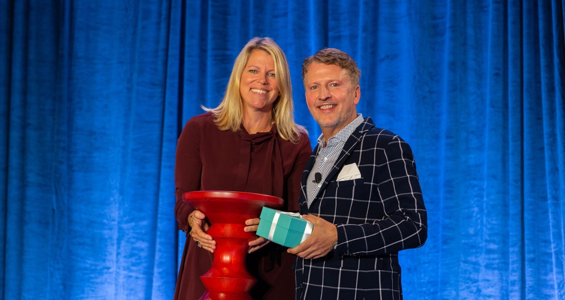Two people stand together on stage, smiling and holding a red pedestal and a blue gift box, with a blue curtain backdrop.