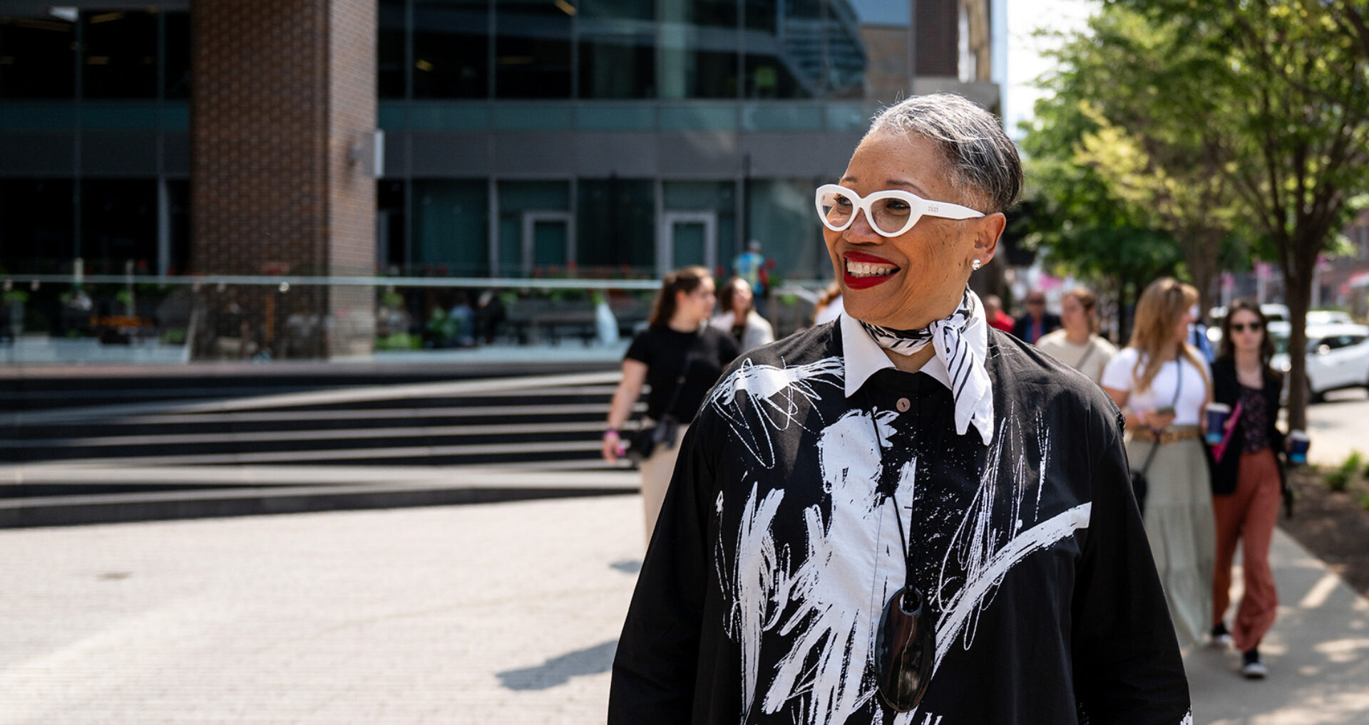 A woman with glasses and a bold black and white outfit smiles while walking, surrounded by a diverse group of people in a sunny urban setting.