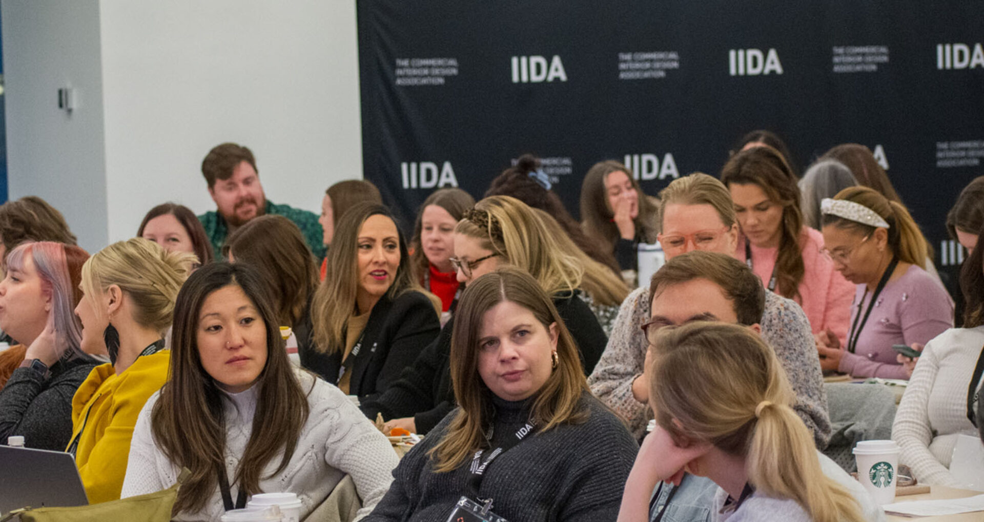 A diverse group of attendees engage in conversation at a professional event, showcasing interest and collaboration amid a backdrop featuring IIDA branding.