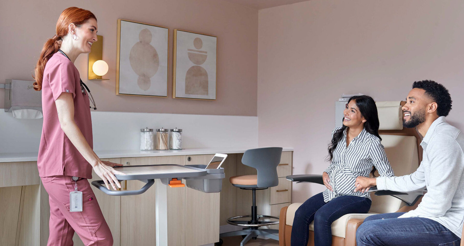 A healthcare provider in scrubs smiles while assisting a pregnant woman and her partner in a warm, welcoming medical office setting.