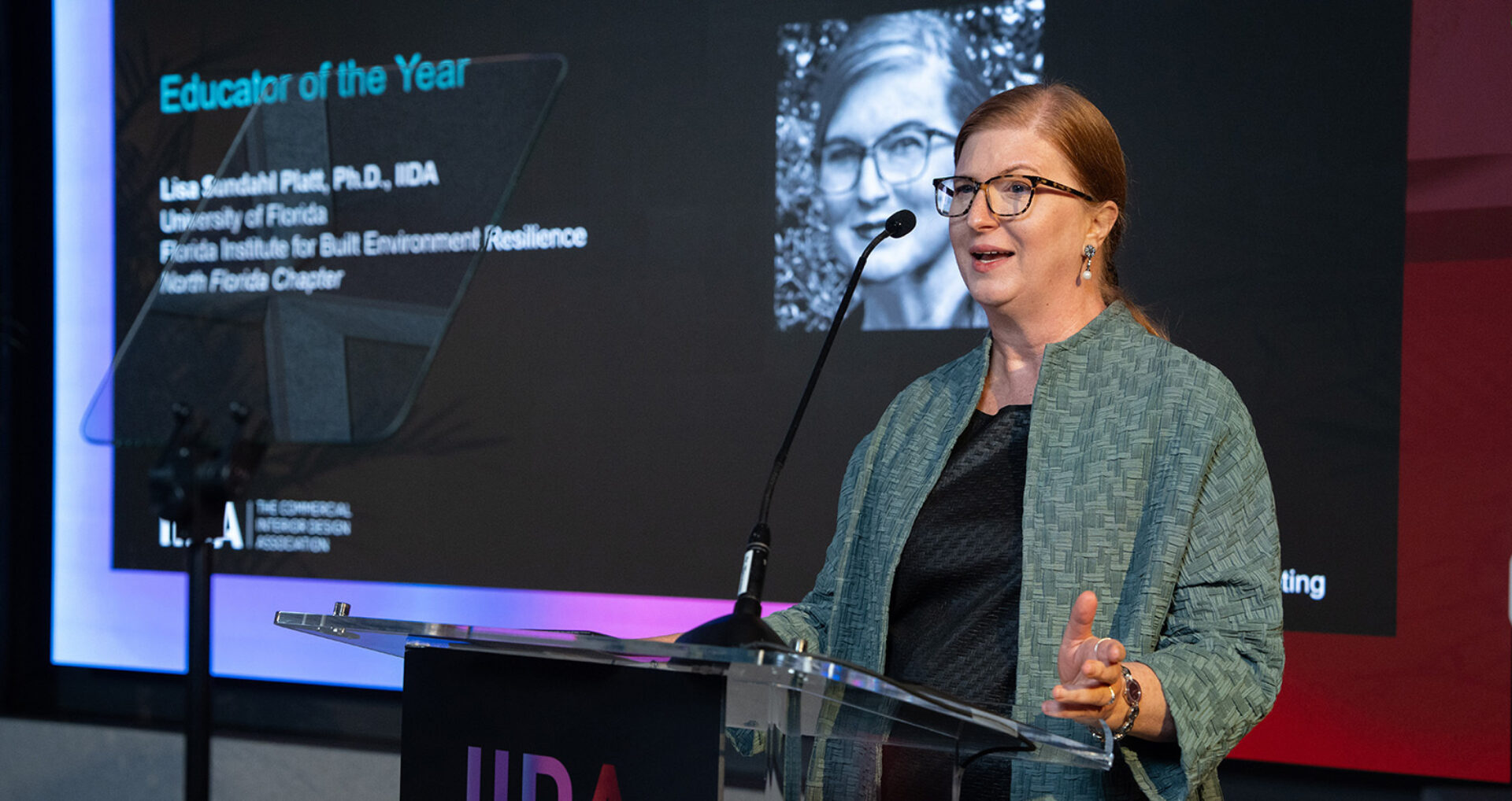 A woman wearing glasses and a green textured jacket speaks at a podium into a microphone, with a slide reading Educator of the Year.