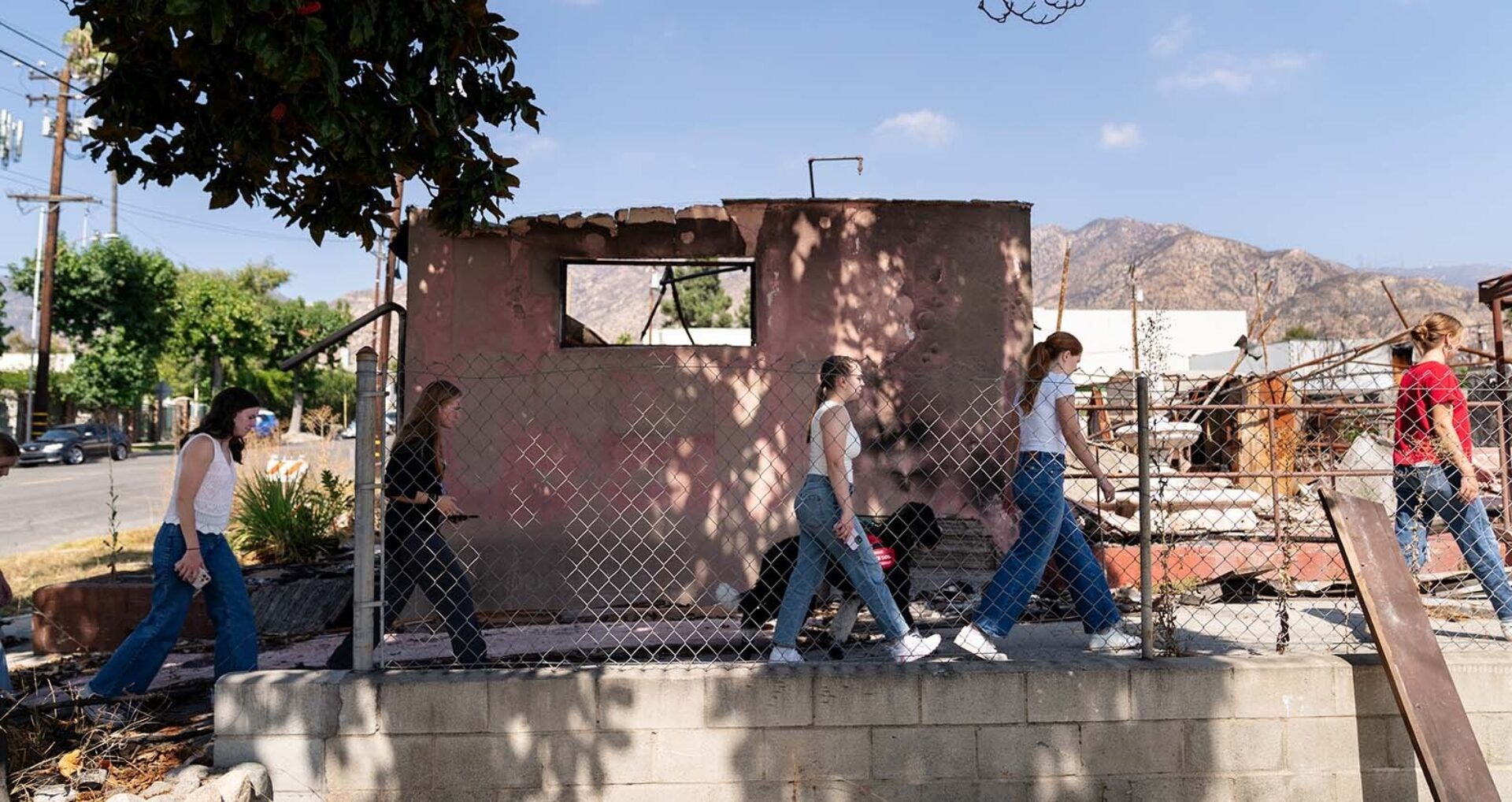 Five young women in jeans walk single file along a chain-link fence past a burned, windowless concrete structure with mountains beyond.