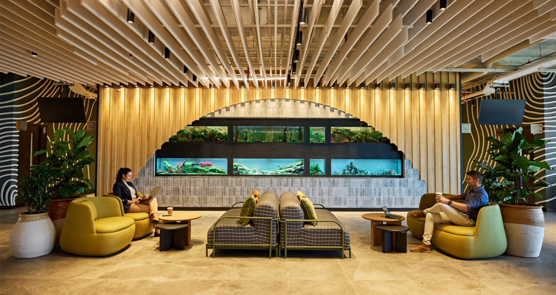 Modern office lounge with a stepped semicircular wall of aquariums, warm wood slats, green lounge chairs, plants and two people working.