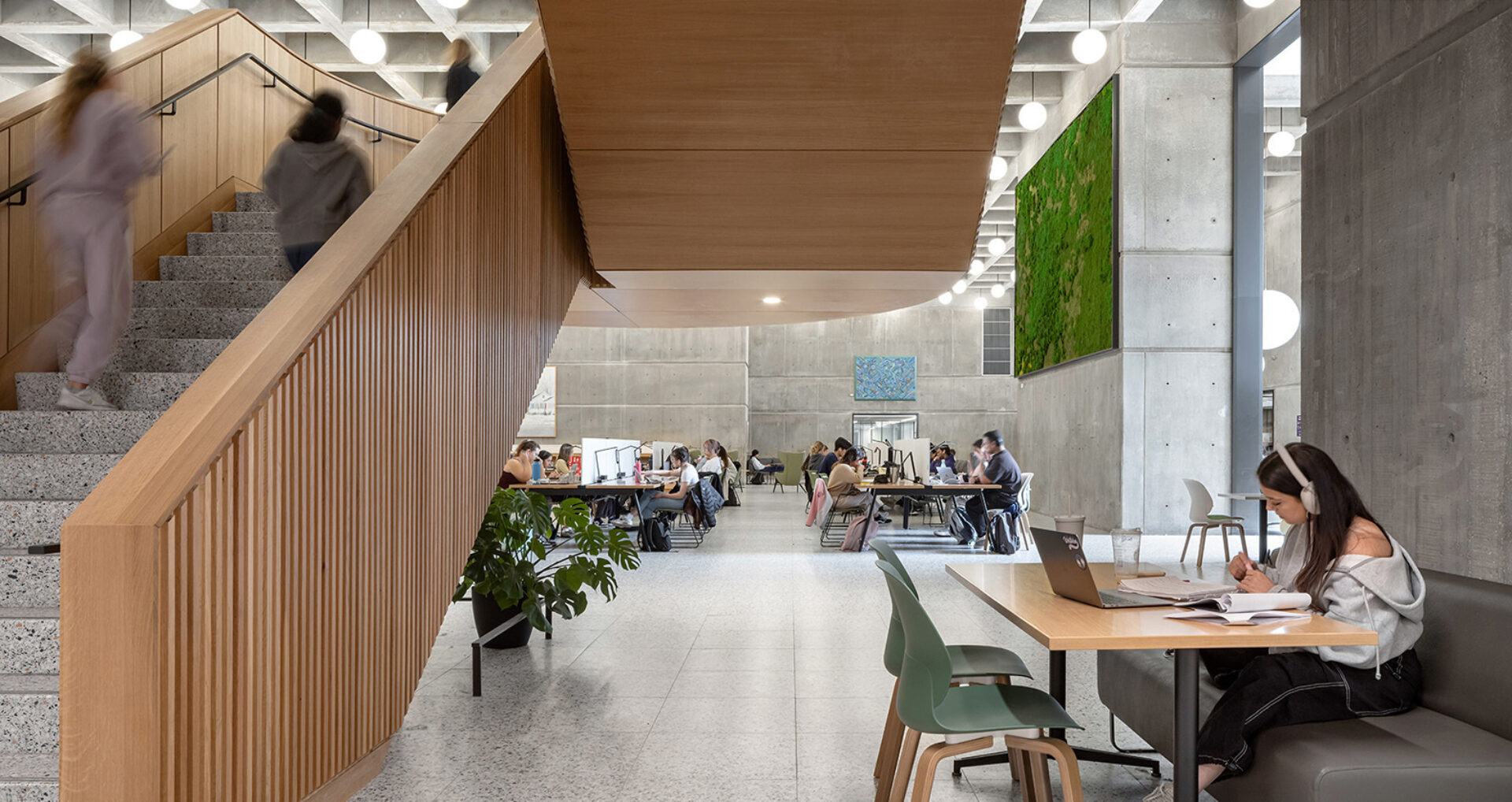 Modern concrete study hall with wooden staircase, students studying at long desks, green moss wall art, and a woman working with headphones.