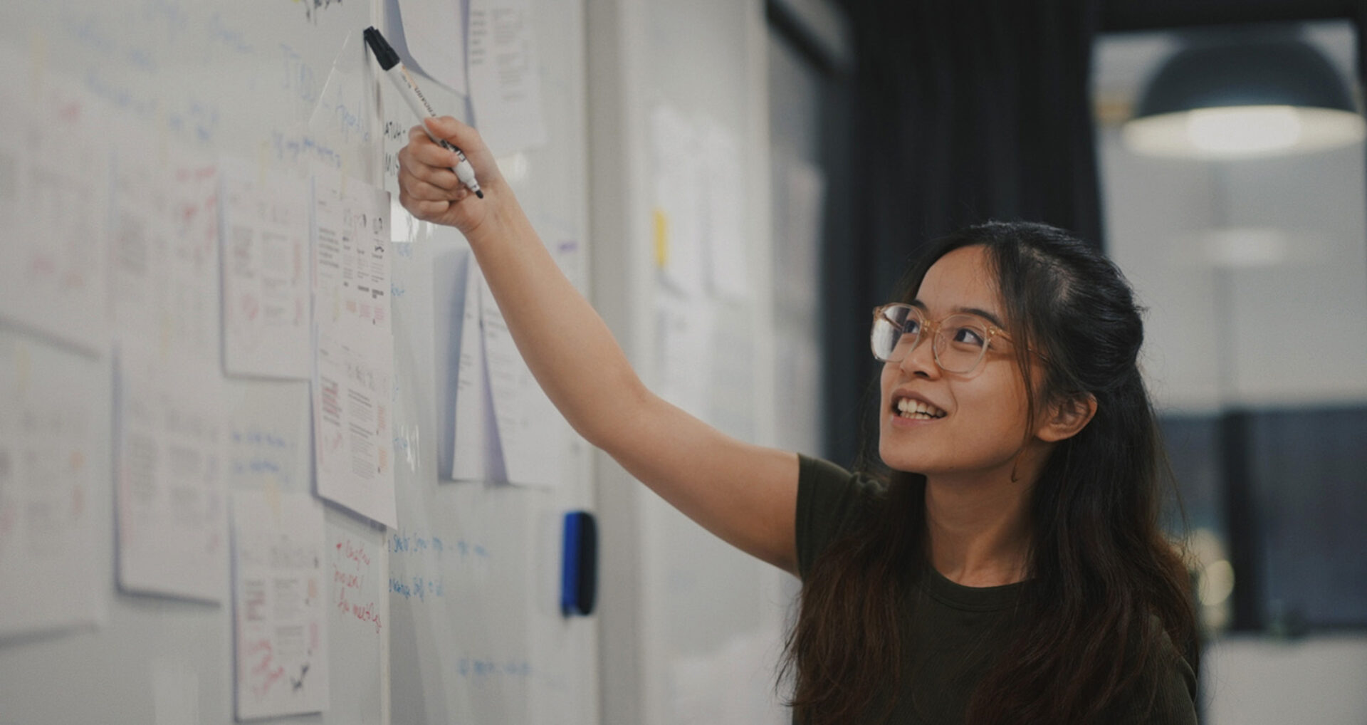 A young woman with glasses engages thoughtfully while writing on a whiteboard filled with notes and papers in a bright, modern workspace.