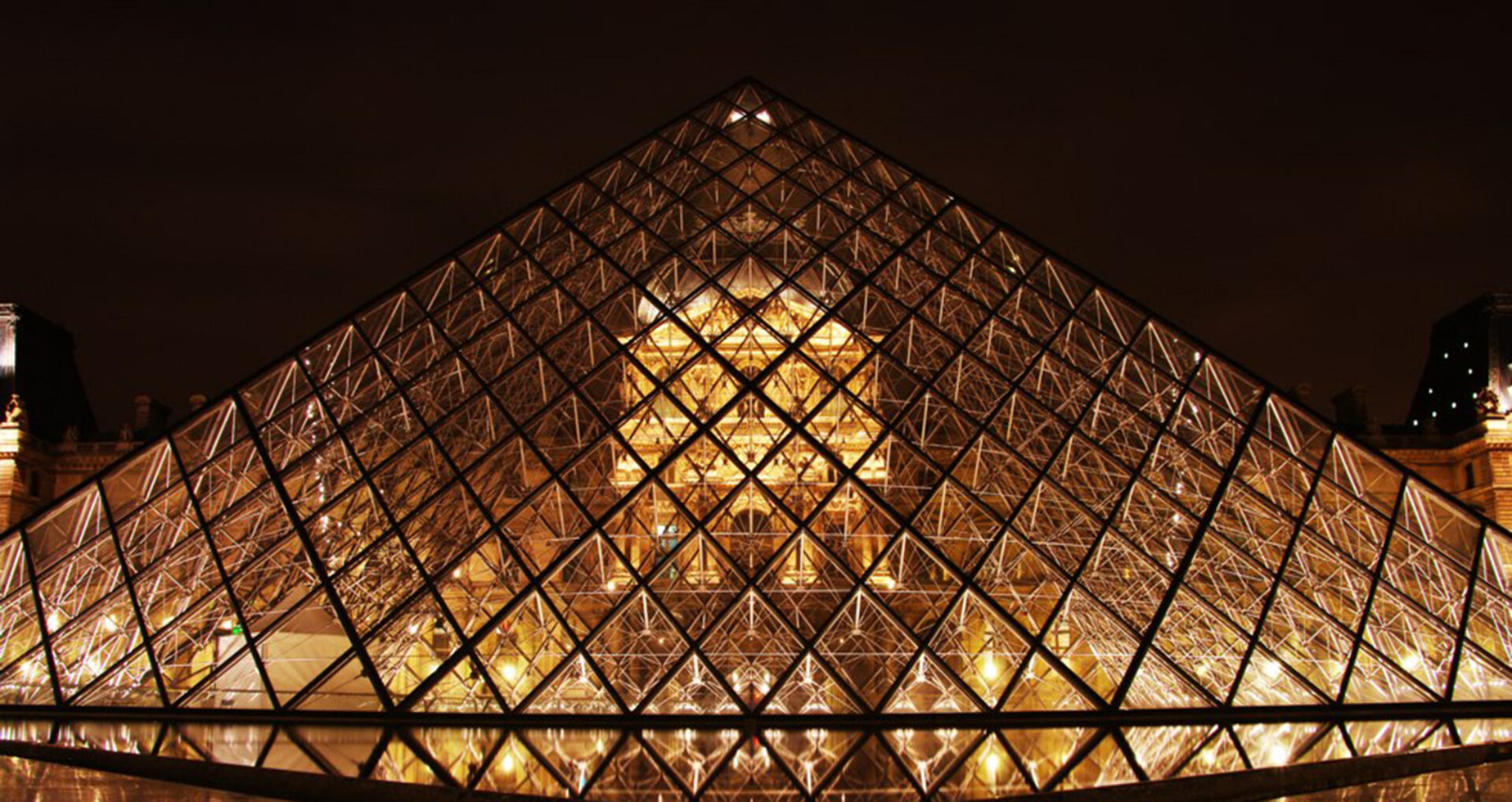 The glass pyramid entrance of the Louvre Museum is illuminated at night, showcasing its geometric design against a dark sky.