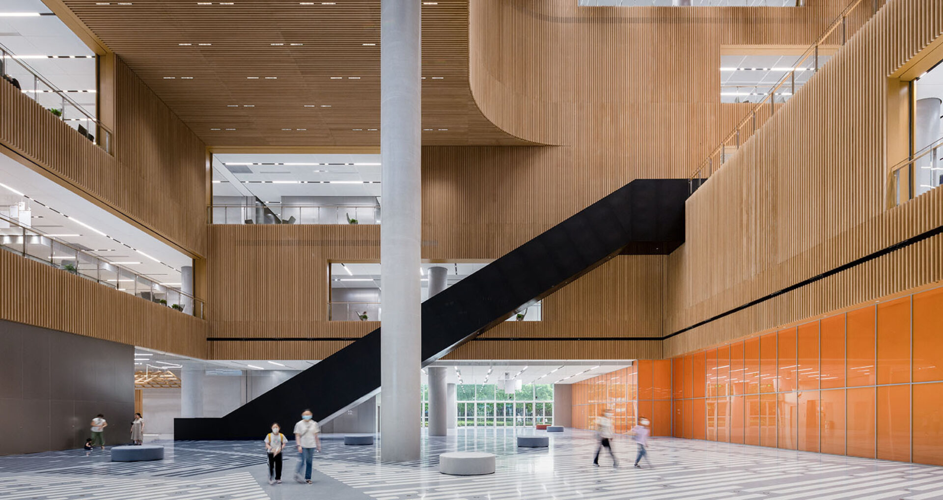A spacious, modern interior showcases wooden walls, large staircases, and people walking across a patterned floor. Natural light floods the area.