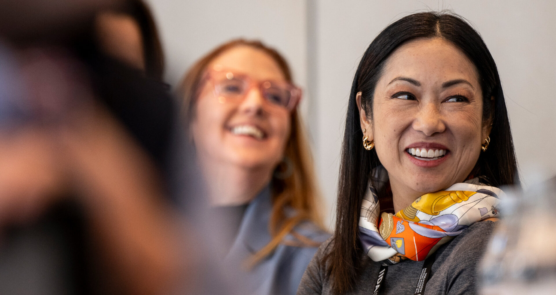 A smiling woman with dark hair and a patterned scarf engages in conversation, with another woman laughing in the background.