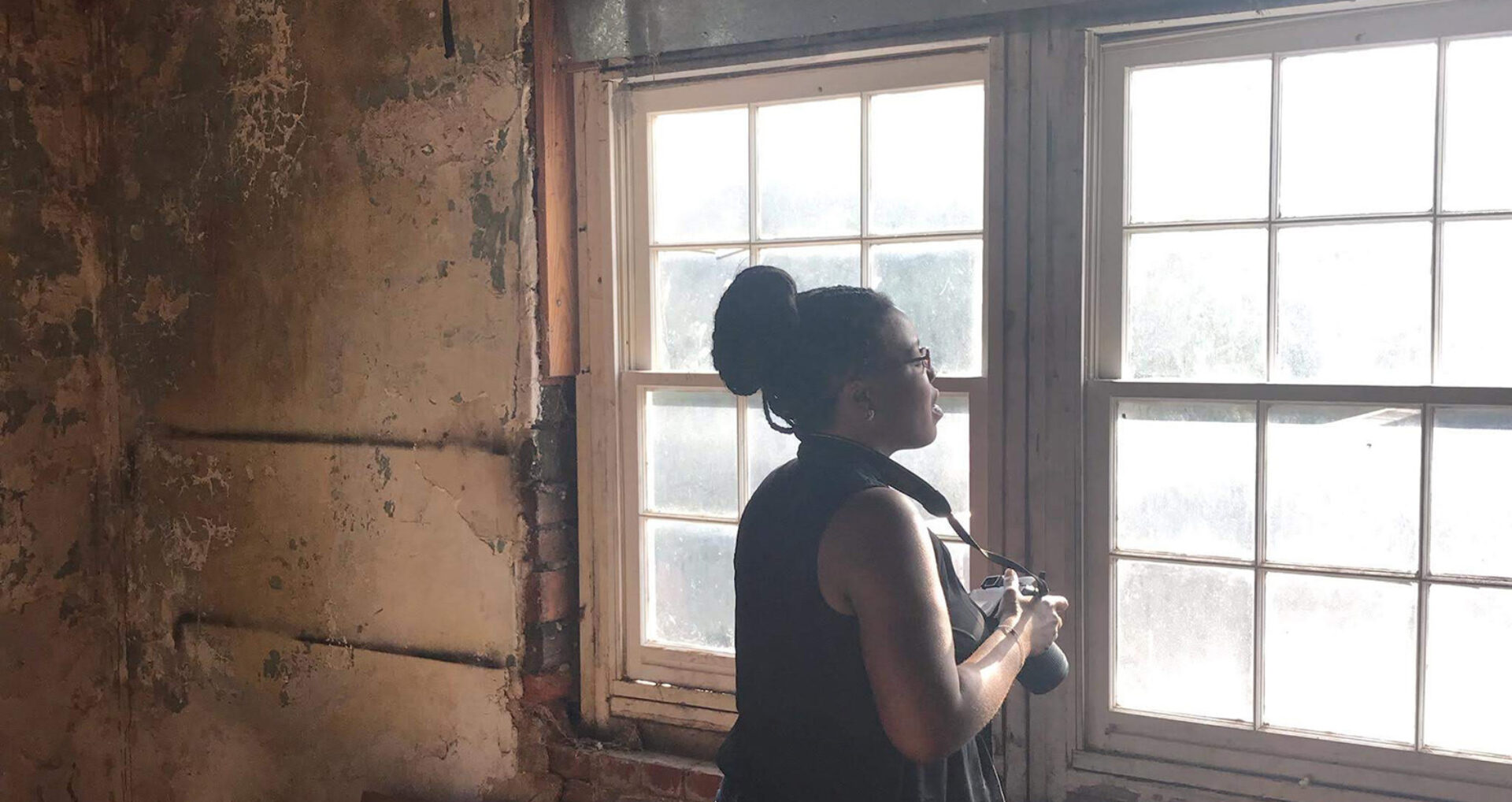 A woman stands by a window in a weathered room, holding a camera and gazing outside, with peeling walls creating a rustic backdrop.
