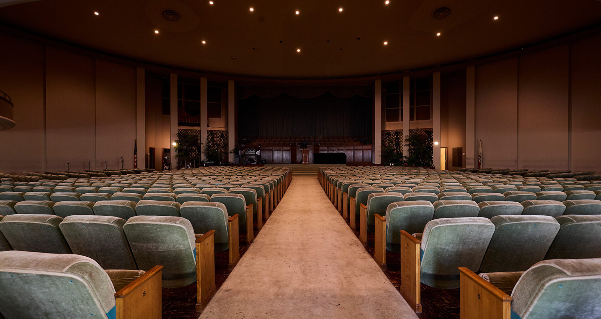 An empty auditorium features rows of green upholstered seats leading to a stage with a dark curtain, illuminated by soft overhead lights.