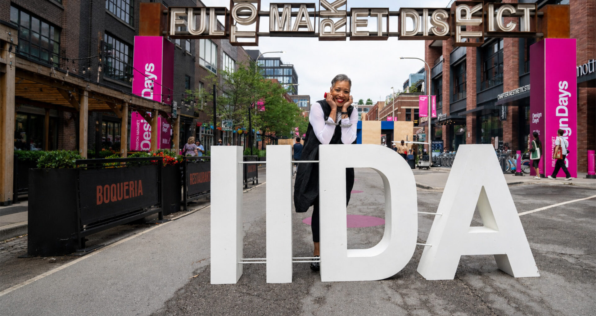 Smiling woman leans on oversized white IIDA letters on a closed Fulton Market street, surrounded by brick shops, pink design banners.