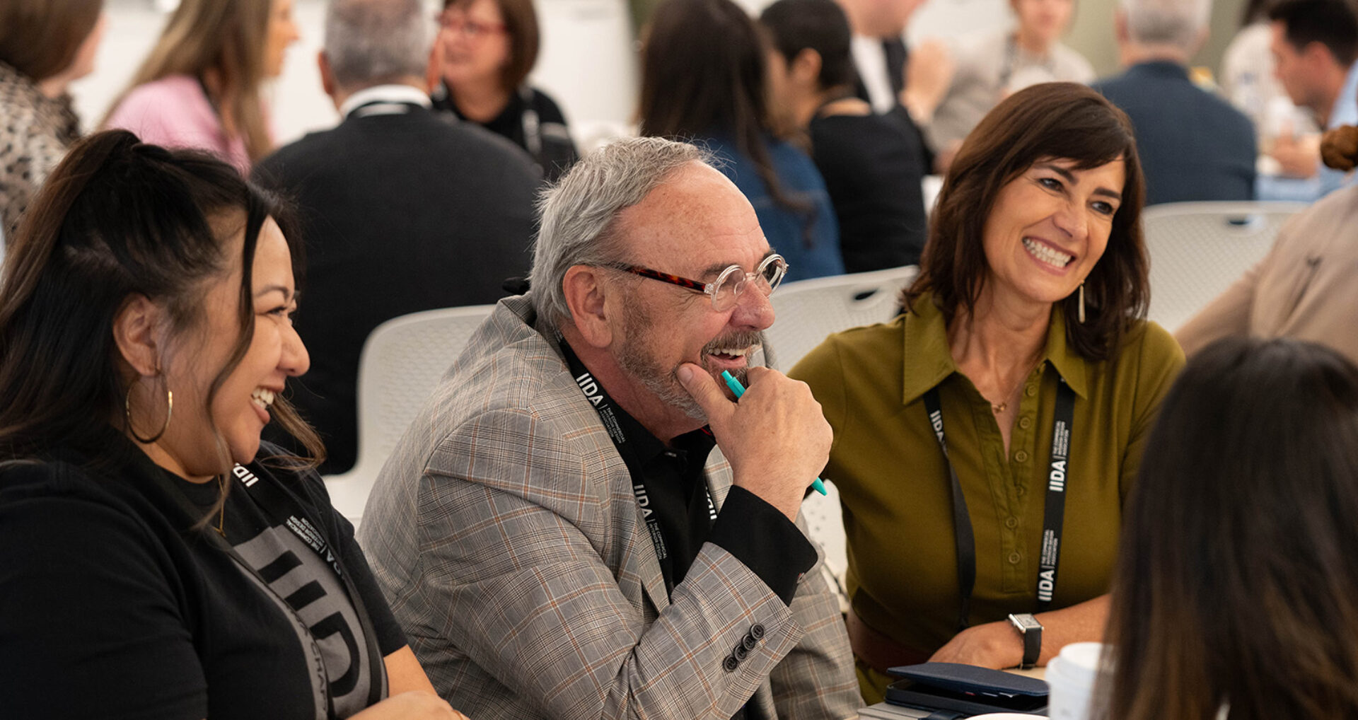 Three people at a conference laugh: a gray-haired man in a plaid blazer, a woman wearing hoops, and a woman in olive with an IIDA lanyard.