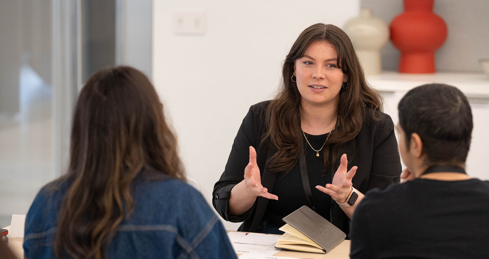 A woman with long hair passionately discusses ideas with two people in a meeting room, gesturing expressively while seated at a table.