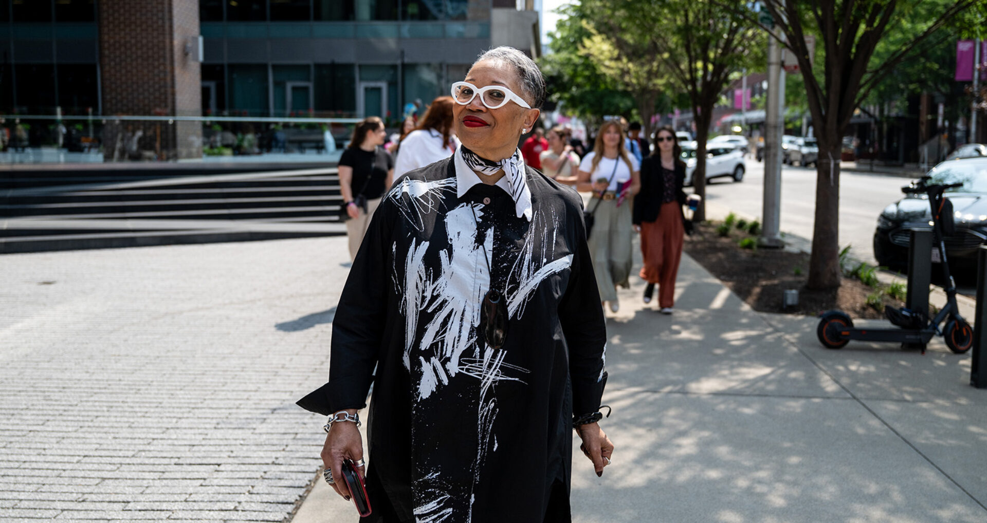 A stylish woman with glasses walks confidently down a city sidewalk, wearing a bold, black and white outfit, surrounded by passersby.