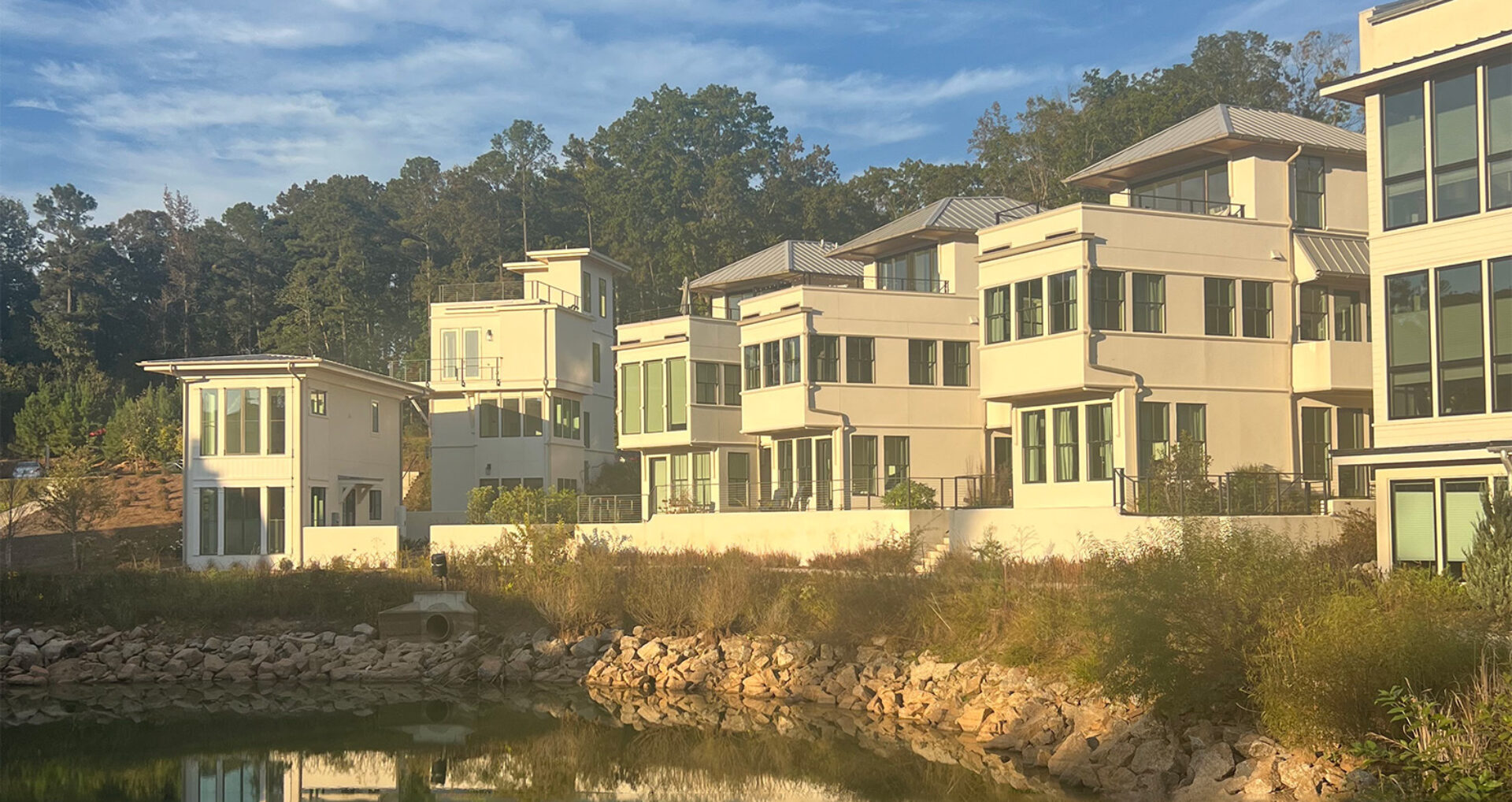 Modern, multi-story homes with large windows sit beside a serene body of water, surrounded by trees and landscaped greenery.