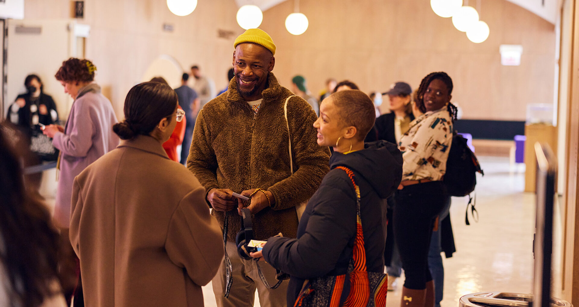 A diverse group of people converse in a lively indoor space, with warm lighting and a modern aesthetic enhancing the social atmosphere.