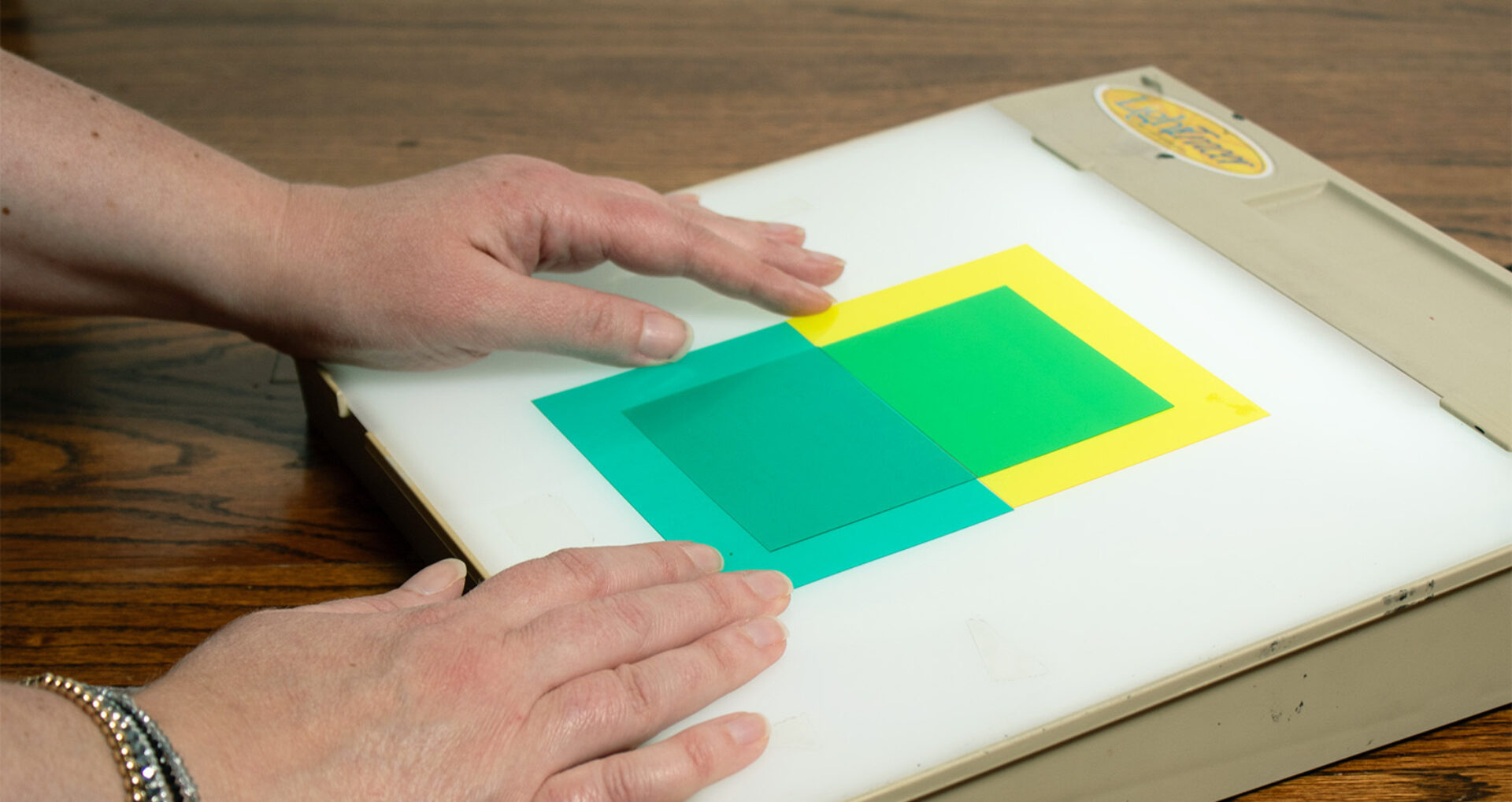 Two hands aligning translucent green and teal square filters overlapping a yellow square on a backlit lightbox atop a wooden table.