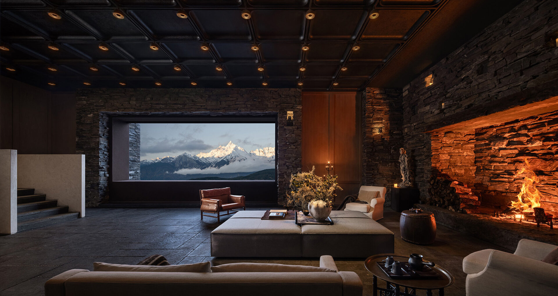 Cozy stone lodge living room with a wide window framing snow-capped peaks, roaring fireplace, leather chair, low ottoman and warm lighting.