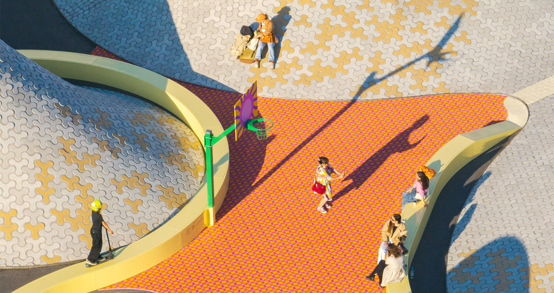 Aerial view of a colorful plaza with curved yellow benches, patterned tiles, a green basketball hoop, a scooter rider, and seated people.