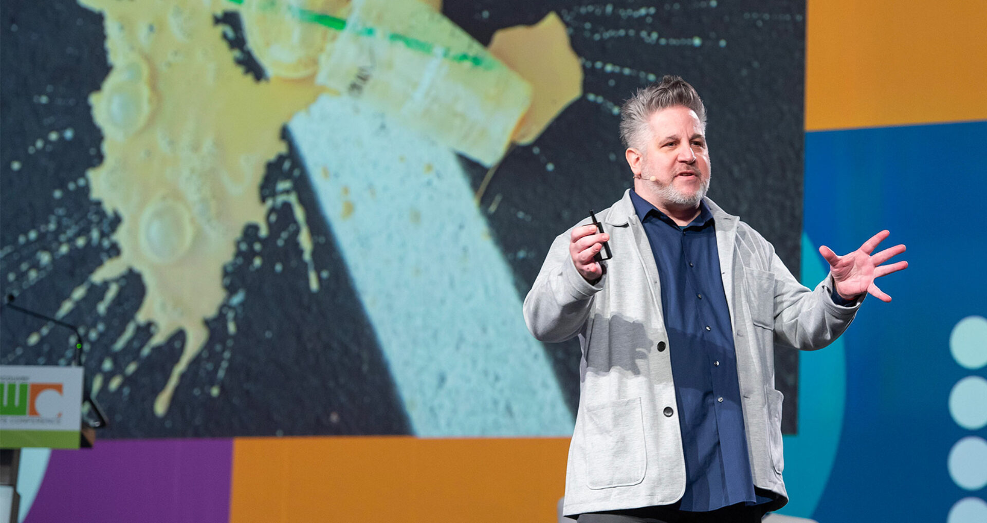 Middle-aged man in a gray jacket and navy shirt speaking on stage with a clicker, gesturing beside a large abstract projection.