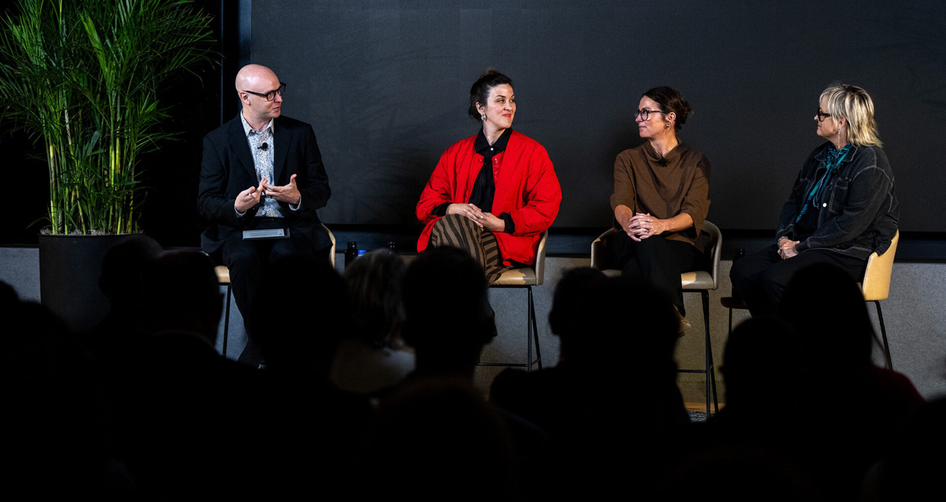 Four panelists on stage: a man in a suit and three women, the center woman wearing a bright red jacket; silhouetted audience in foreground.
