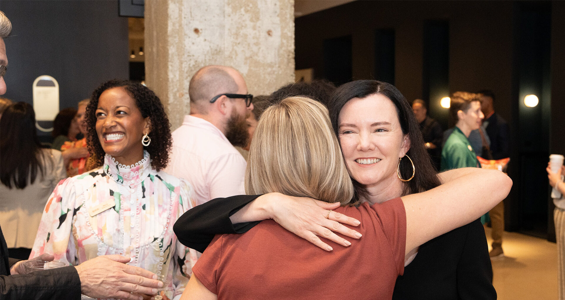 Black-haired woman with gold hoop earring hugs a blonde from behind at a bustling networking event while a woman in a floral blouse smiles.