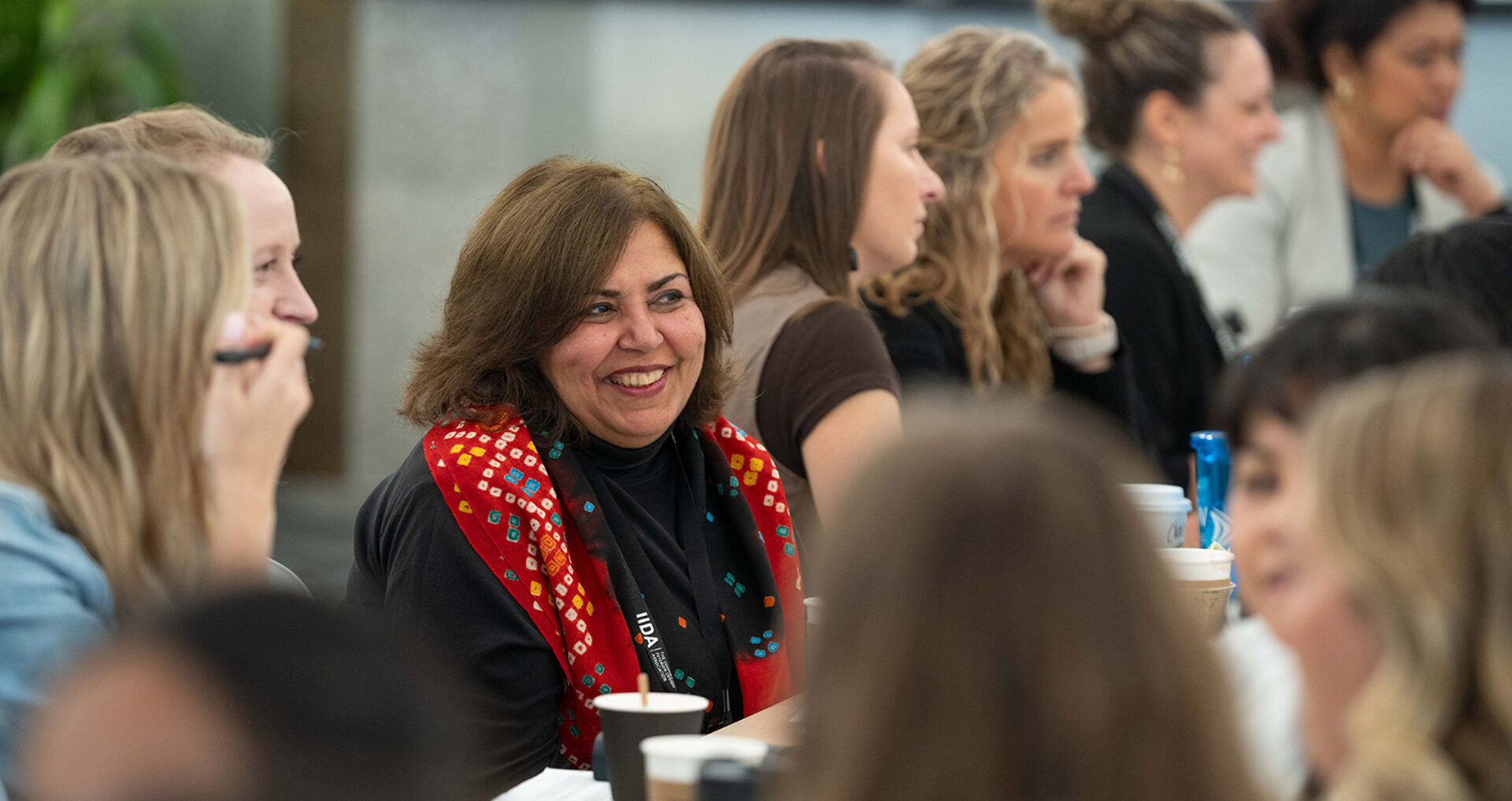 A diverse group of women engages in conversation at a round table, with one woman in a vibrant scarf smiling warmly.