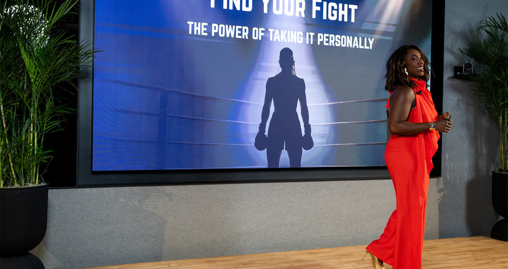 Smiling Black presenter in a red jumpsuit stands on stage beside a large screen showing a silhouetted female boxer and slogan.