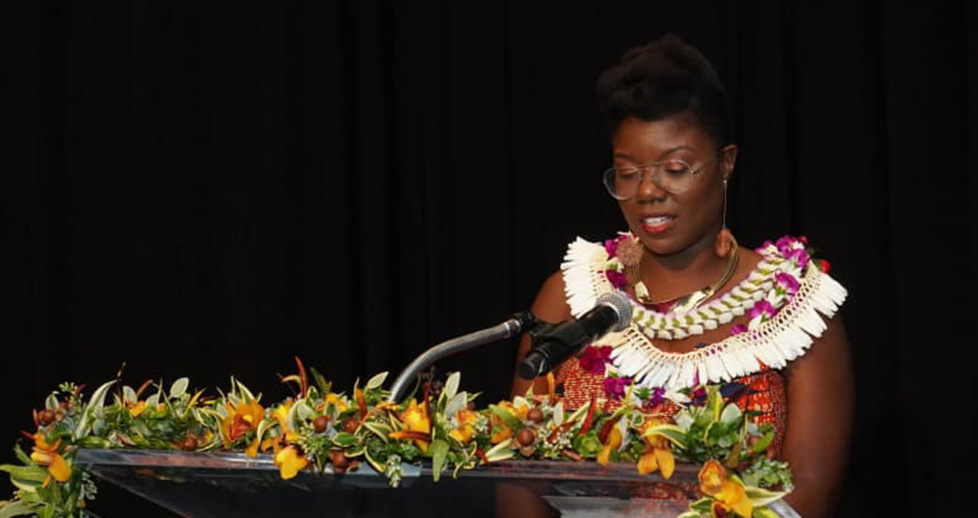 A woman stands at a podium adorned with vibrant floral arrangements, wearing traditional attire and a decorative necklace, speaking intently.