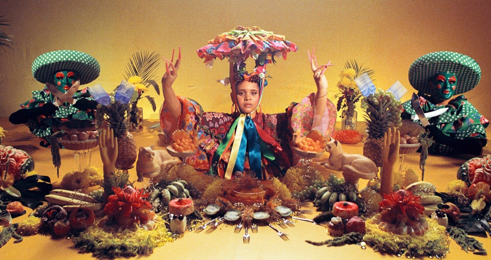 A colorful altar scene featuring a child in vibrant attire, surrounded by fruit, decorative figures, and two masked figures on either side.