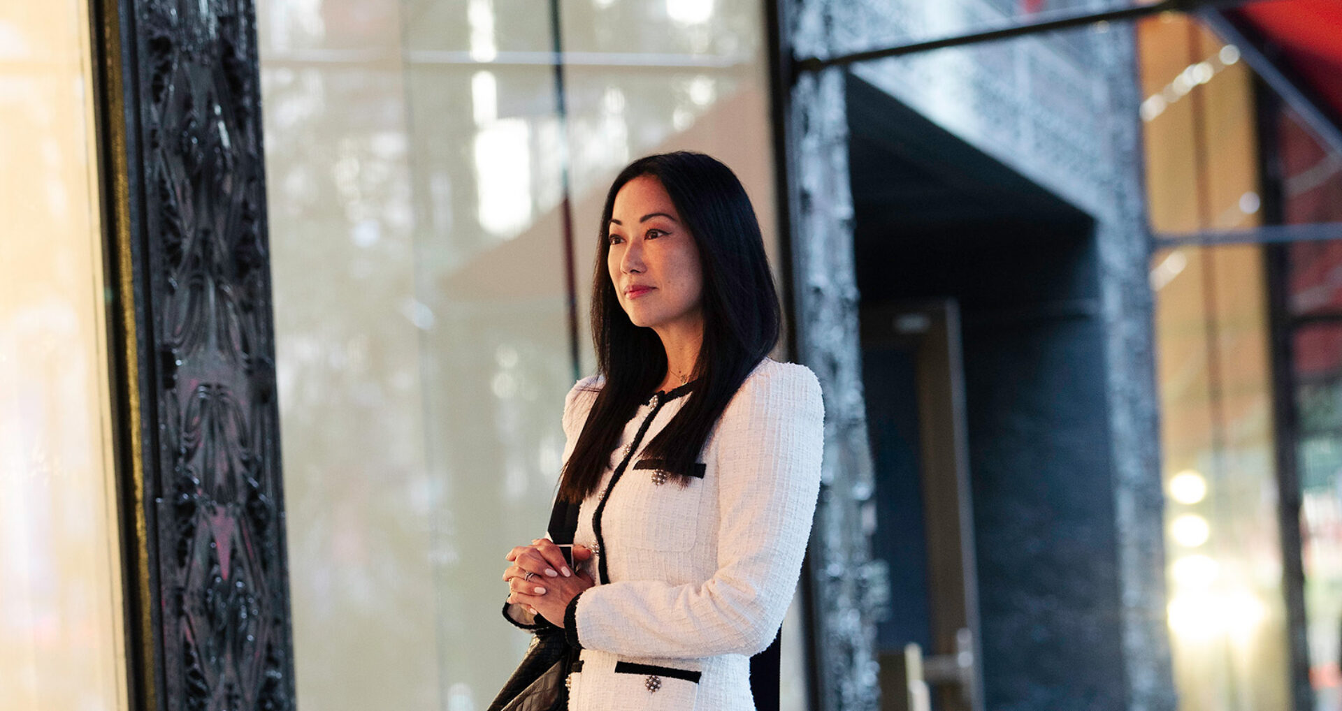A woman with long black hair stands outside a boutique, dressed in a stylish white blazer with black accents, looking contemplative.