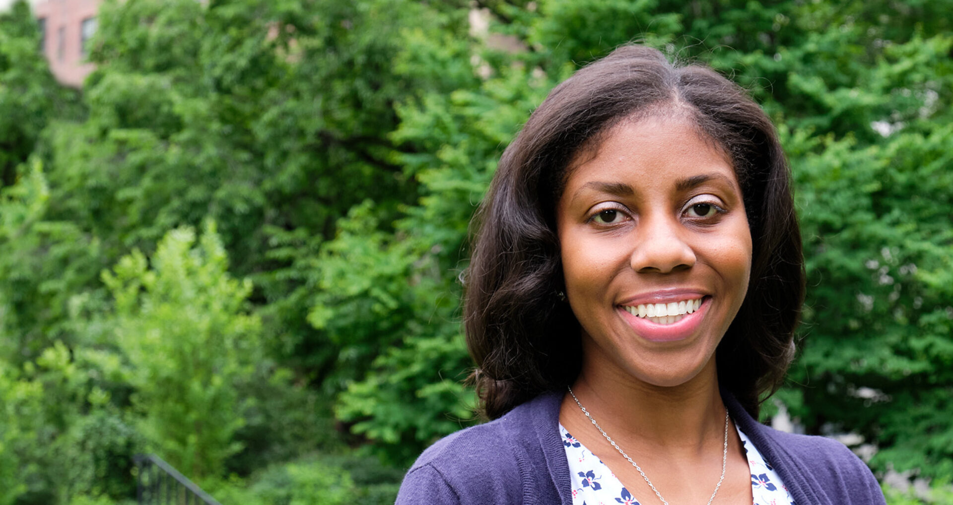A woman with long, dark hair smiles warmly, standing outdoors against a backdrop of lush green trees, wearing a floral blouse and cardigan.