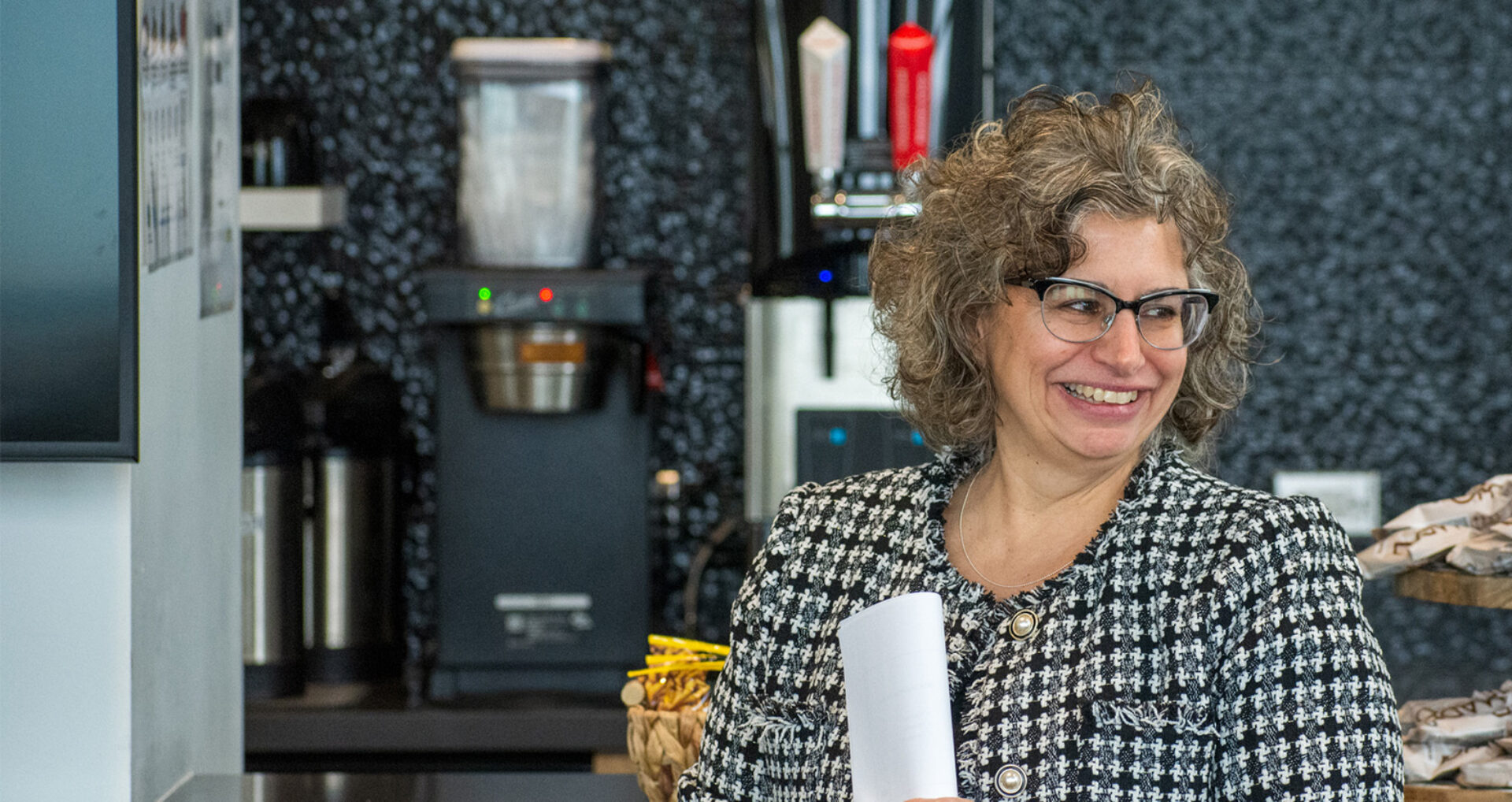 Smiling middle-aged woman with curly gray hair and glasses wearing a black-and-white houndstooth jacket, holding papers by a coffee station.