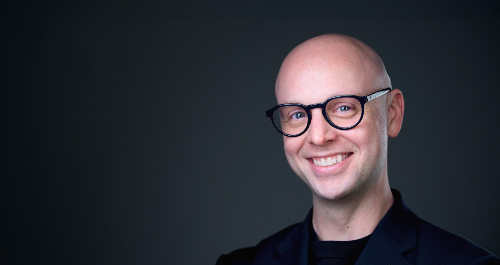 Smiling bald man with round black glasses wearing a dark blazer and black shirt, facing camera against a smooth charcoal-gray background.