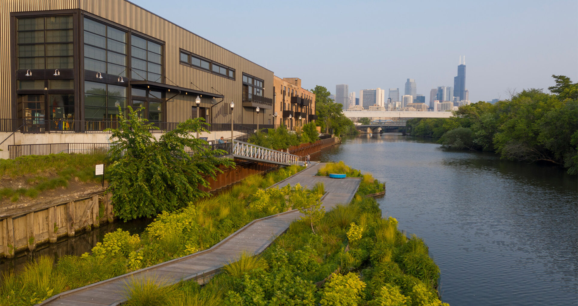 Urban landscape featuring a river bank with lush greenery, a wooden walkway, and modern buildings against a city skyline.