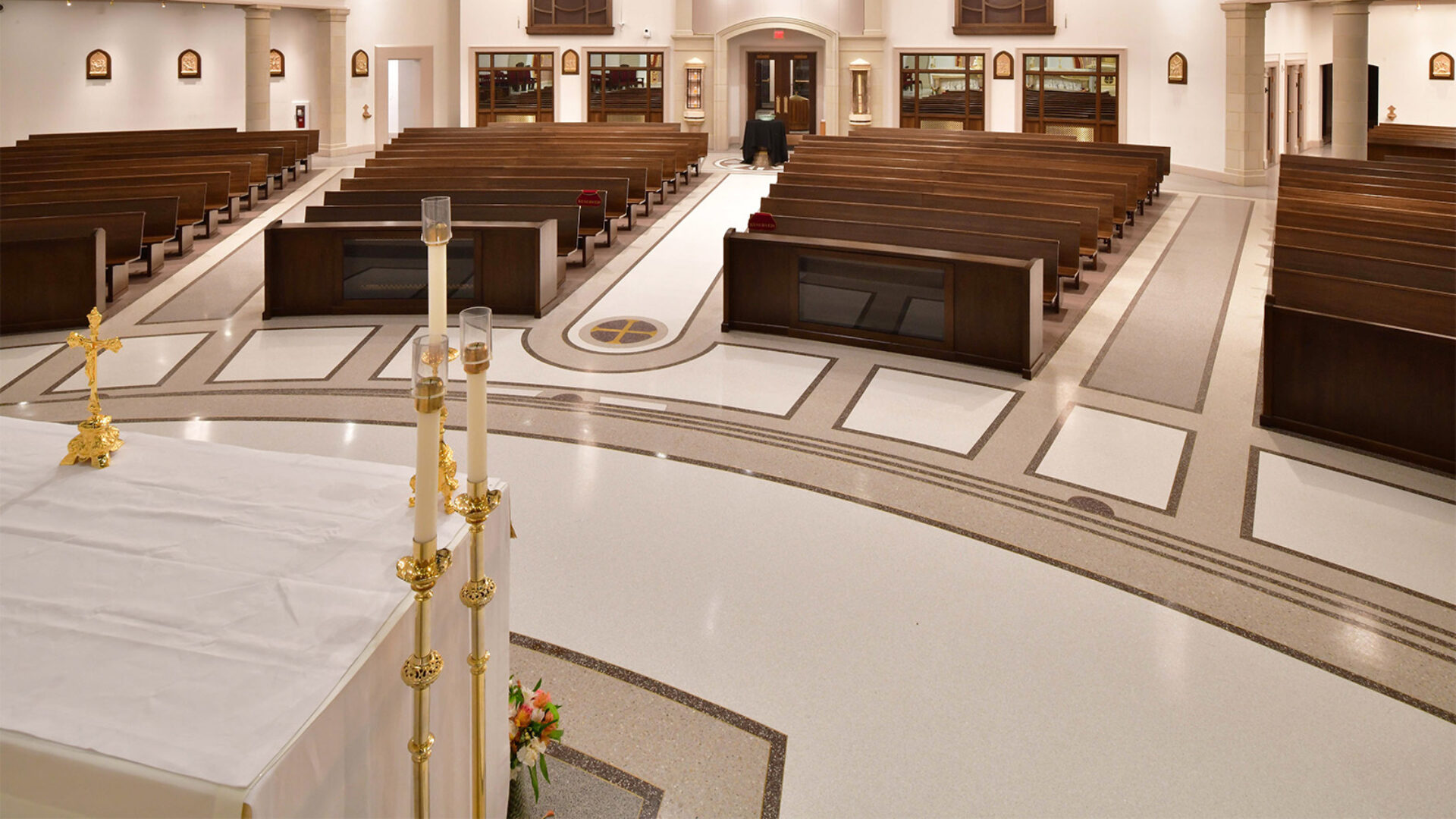 Wide nave of a modern church seen from the altar, showing polished patterned floor, rows of dark wooden pews, central aisle and candles.