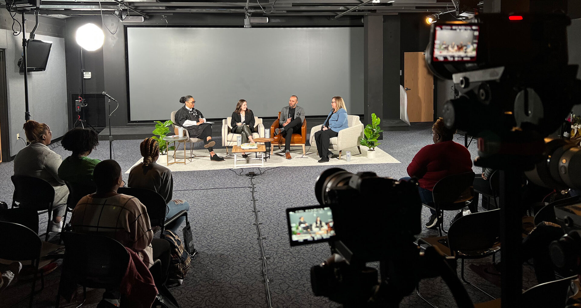A panel discussion featuring four speakers on stage, with an audience seated in front, surrounded by cameras and studio lighting.