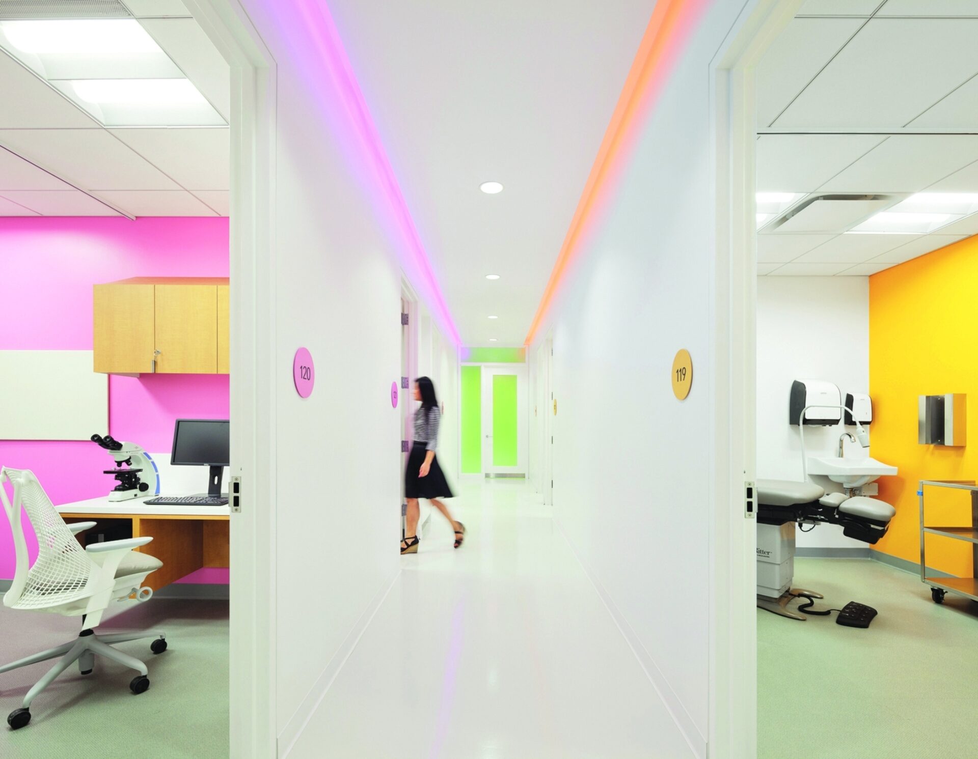 Brightly lit hallway in a medical facility, featuring colorful walls and numbered room doors, with a person walking towards a green door.