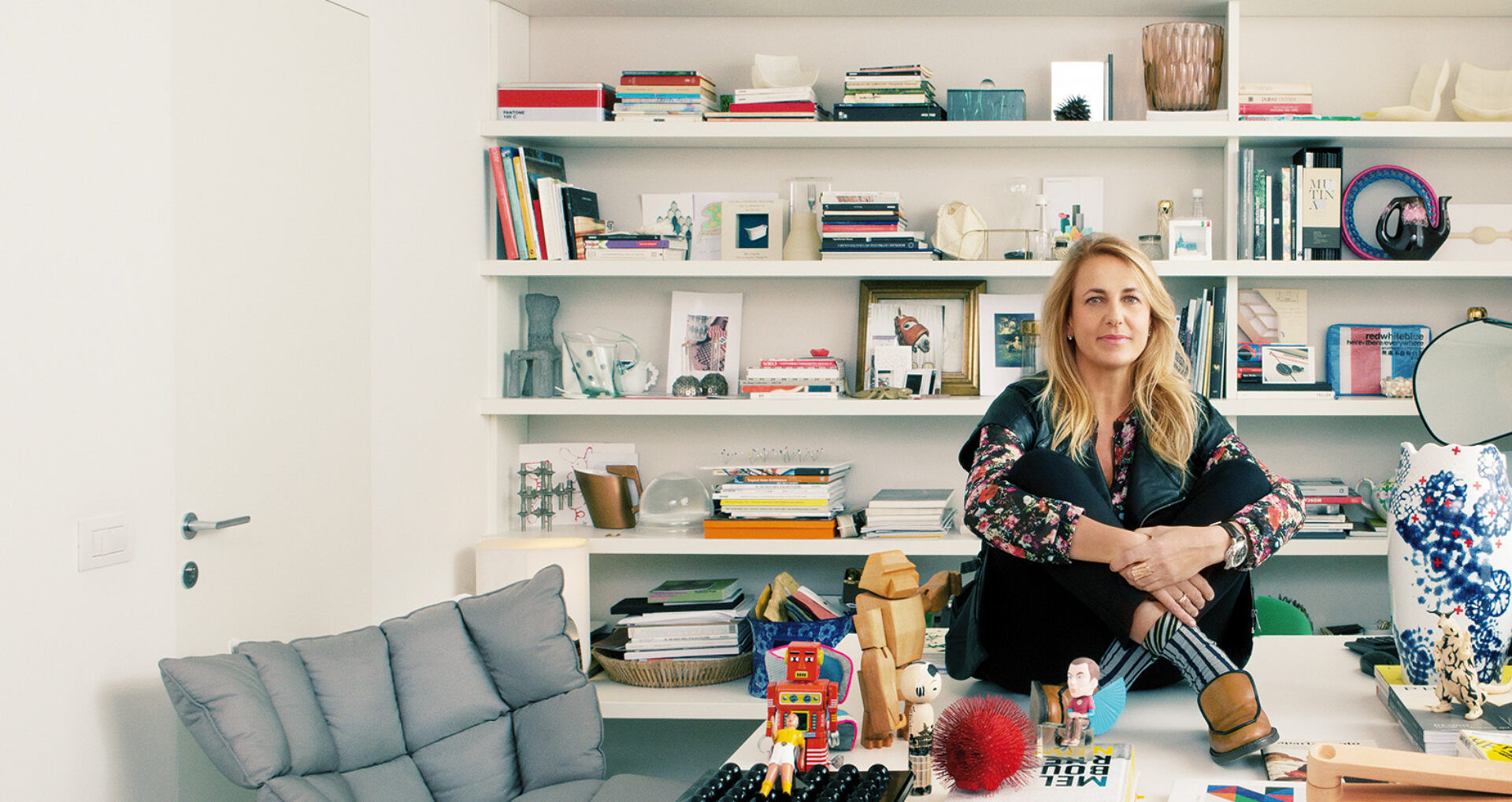A woman sits cross-legged on a desk surrounded by colorful books, toys, and decorative items, with a modern, stylish backdrop.