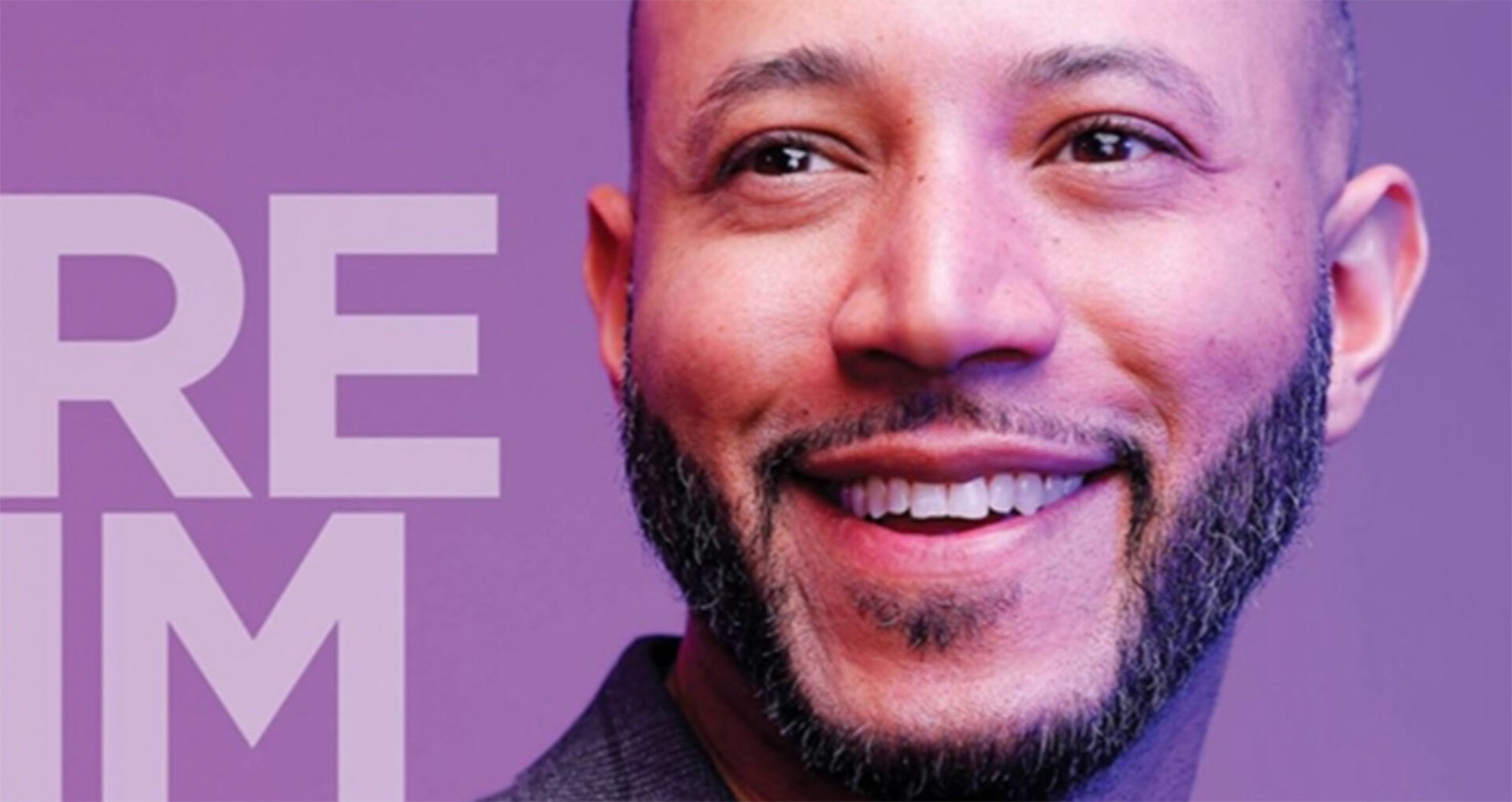 A man with a short beard smiles warmly against a soft purple background, conveying a sense of approachability and confidence.