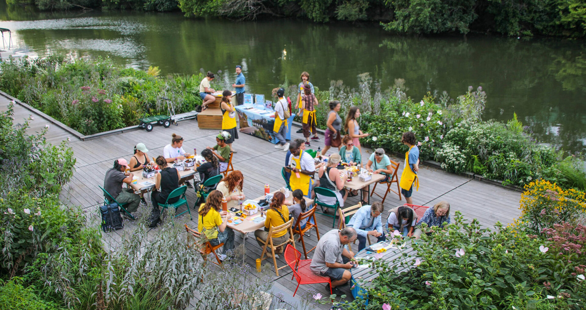 A vibrant gathering by a river features diverse individuals enjoying a meal at tables surrounded by lush greenery and flowers.