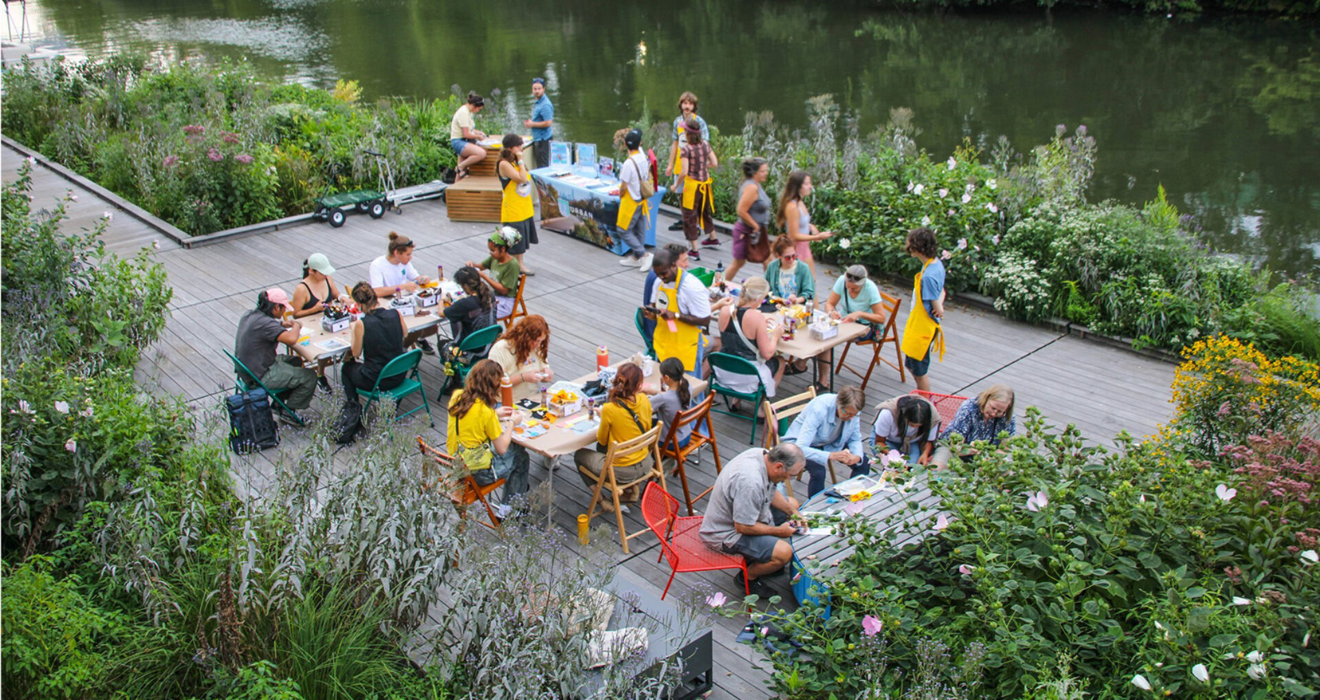 Groups of people sit at tables on a wooden deck by a river, engaging in activities surrounded by vibrant greenery and flowers.