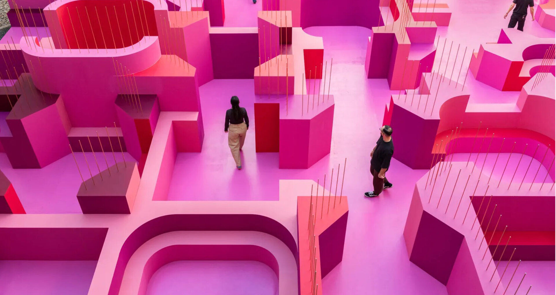 Three people wander a large hot-pink maze of platforms and curved walls studded with thin vertical gold rods, seen from above in soft light.