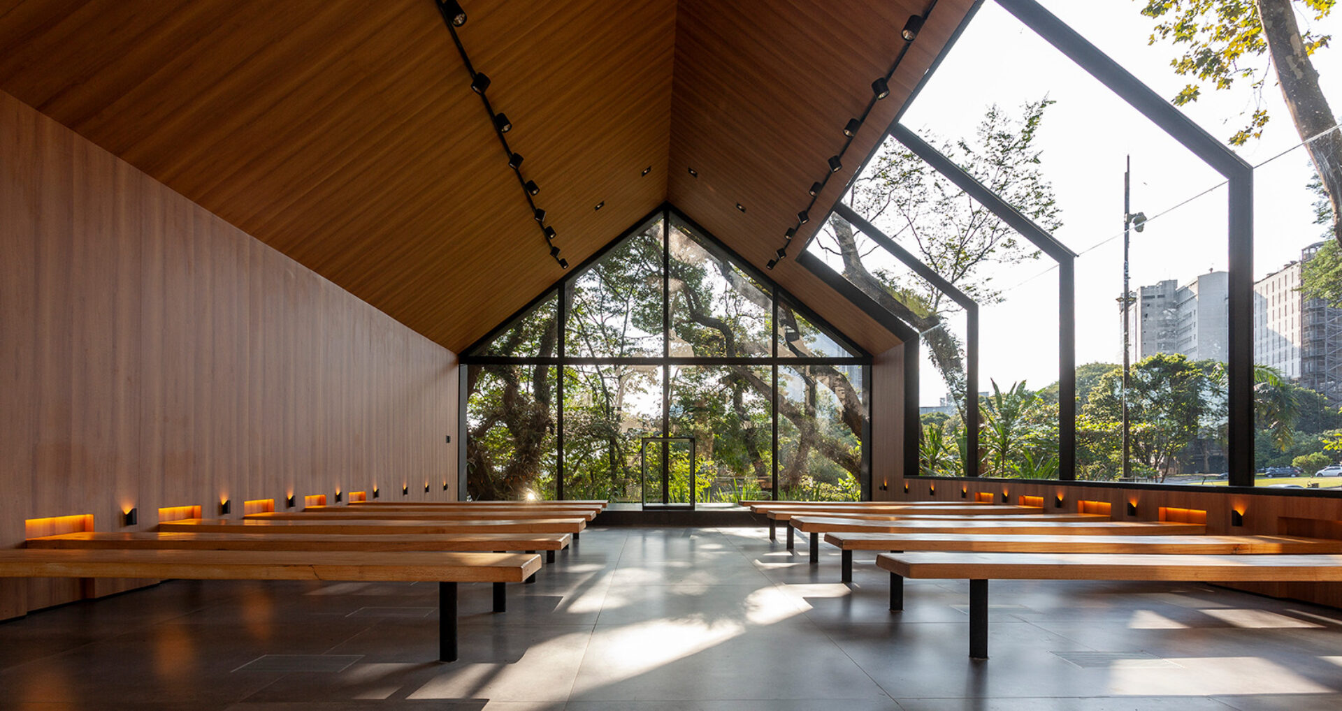 Sunlit modern chapel interior with wooden vaulted ceiling, rows of simple wooden benches, floor-to-ceiling glass revealing trees and city.