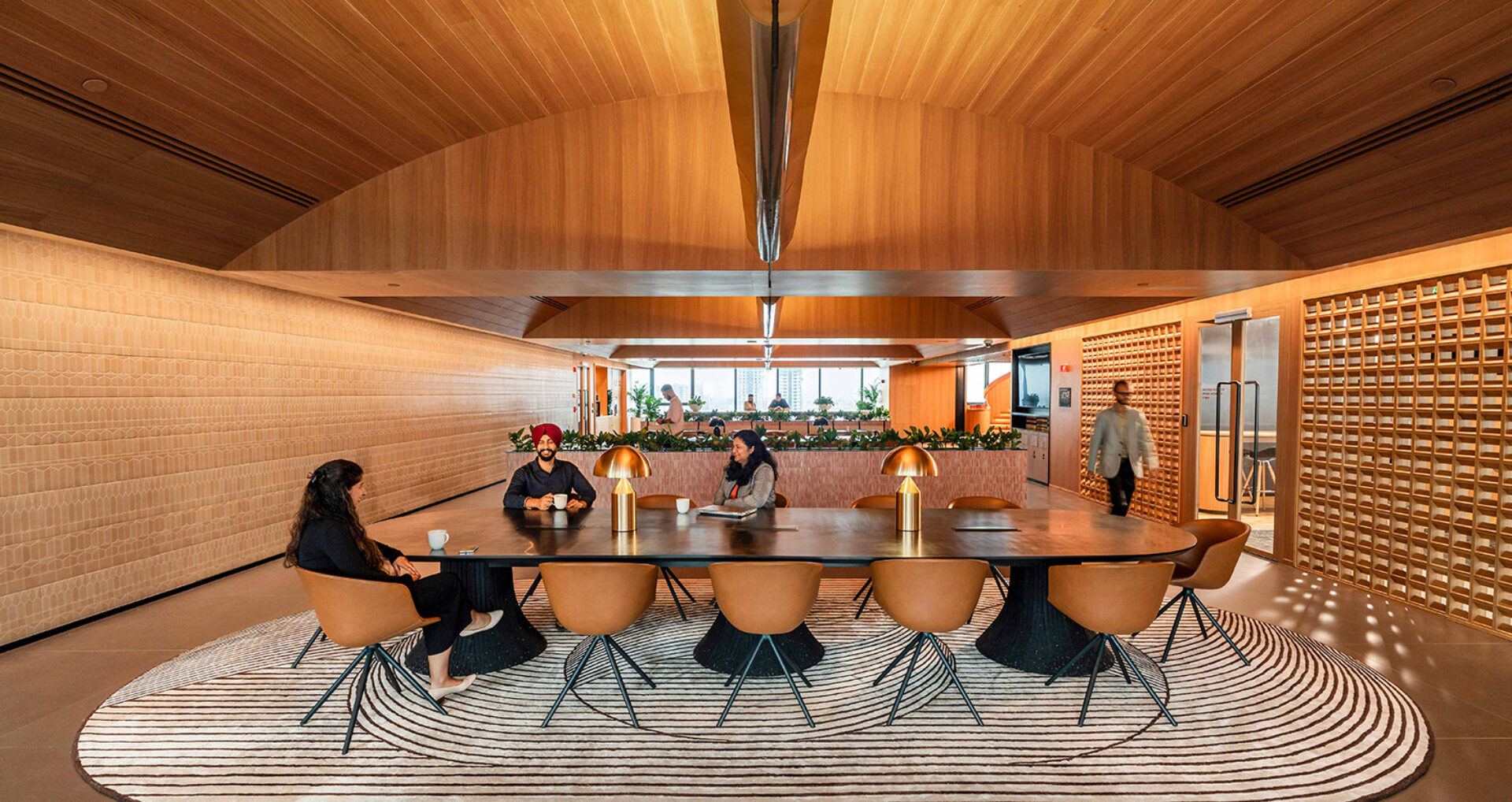 A modern conference room features a wooden ceiling, a large table, stylish chairs, and plants, with people engaged in conversation.