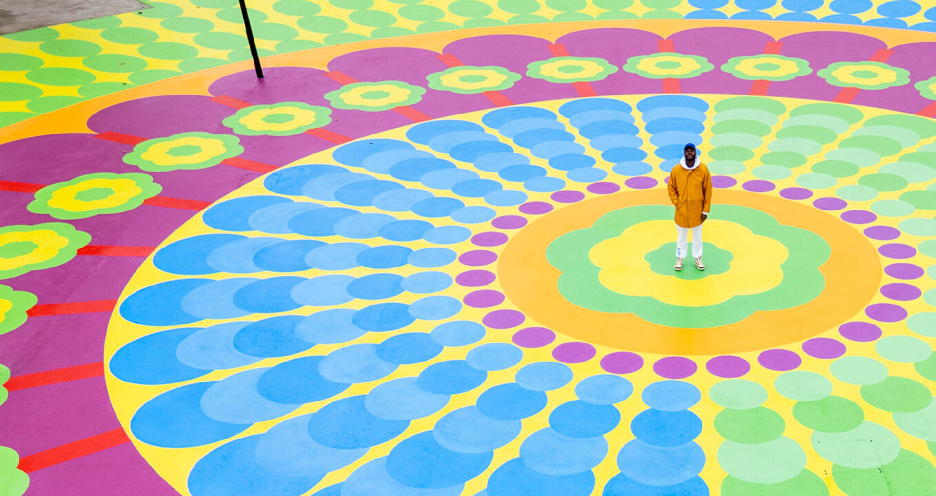 Man in a mustard coat, white pants and cap stands at the center of a giant colorful circular mural of blue, green, purple and yellow.